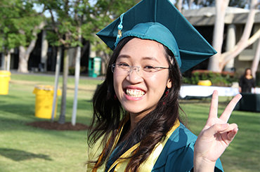 Young woman in graduation cap and gown, smiling at the camera and making a peace sign gesture